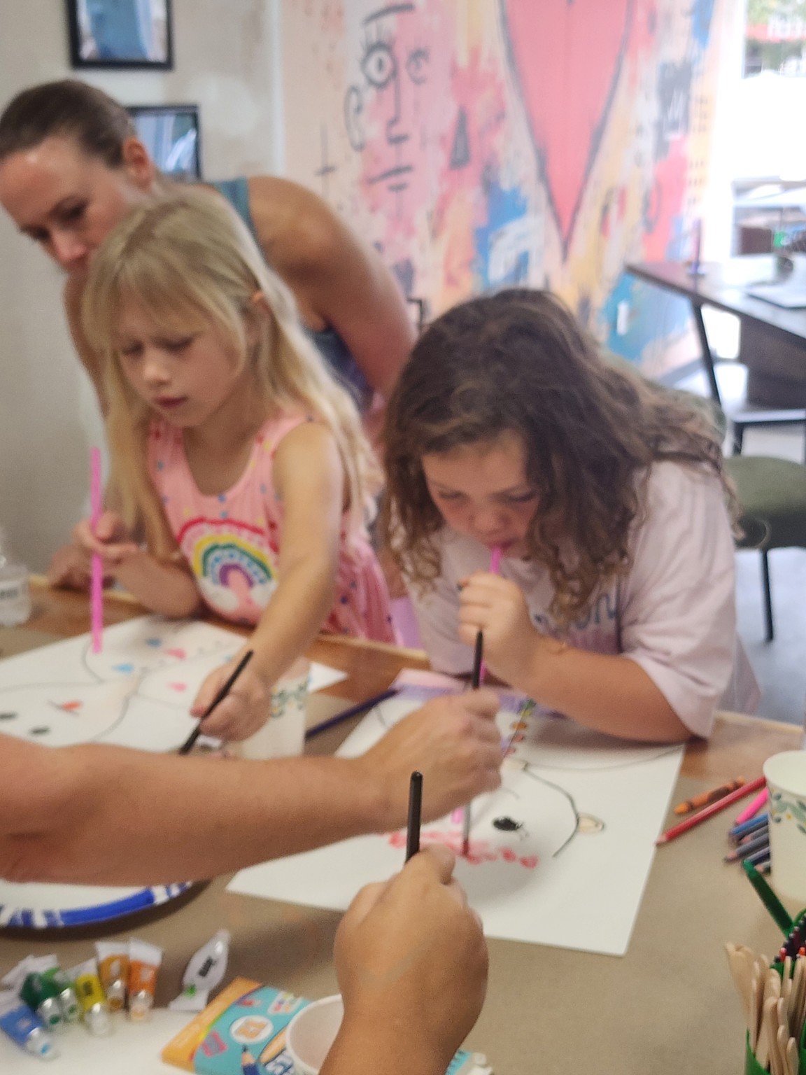 Two children painting at a craft table while an adult leans in to help