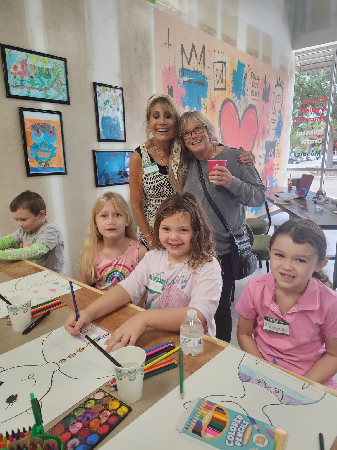 Smiling kids coloring at a table while two adults look on near a painted heart mural