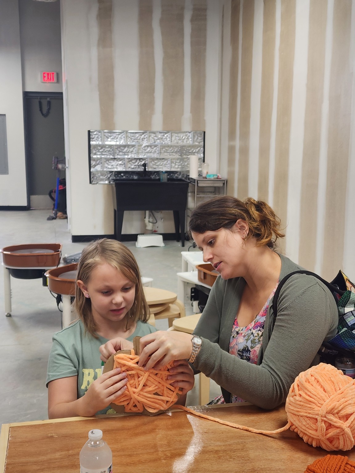 An adult helping a child wrap an orange yarn pumpkin at a craft table